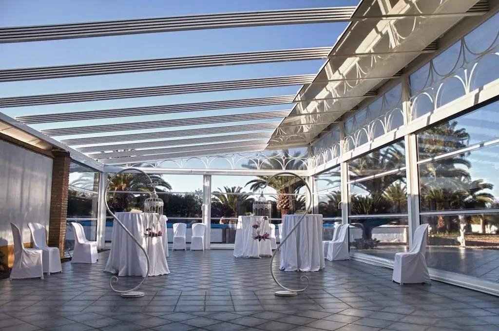 La terraza acristalada con techo de vigas blancas y mesas altas, abierta a las palmeras